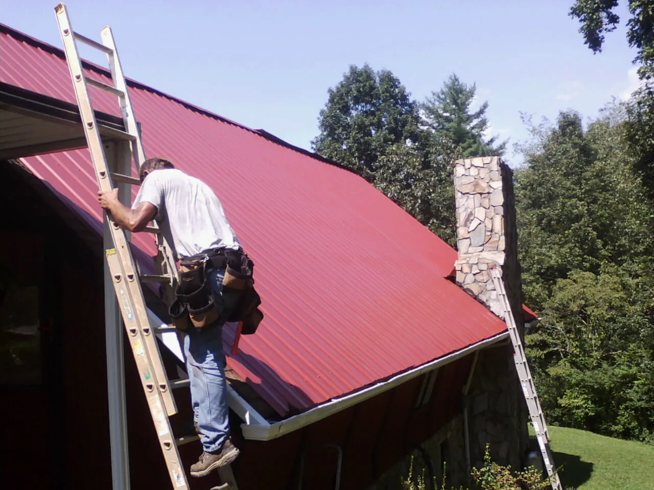 Roofer on a ladder beside a red metal roof and stone chimney — Soffit & Fascia Repair by Roofing Brothers in Deep River